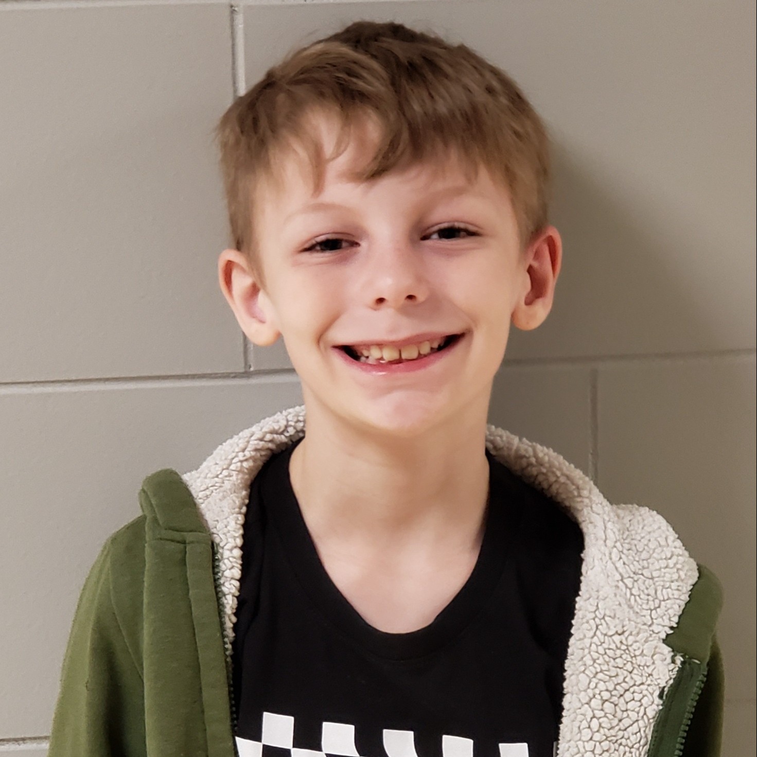 smirking boy with blonde hair wearing a black t-shirt under a green jacket