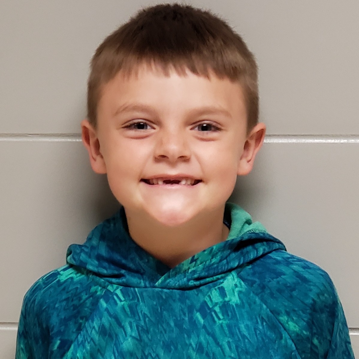 smiling young boy with light brown hair missing front teeth wearing a teal hoodie