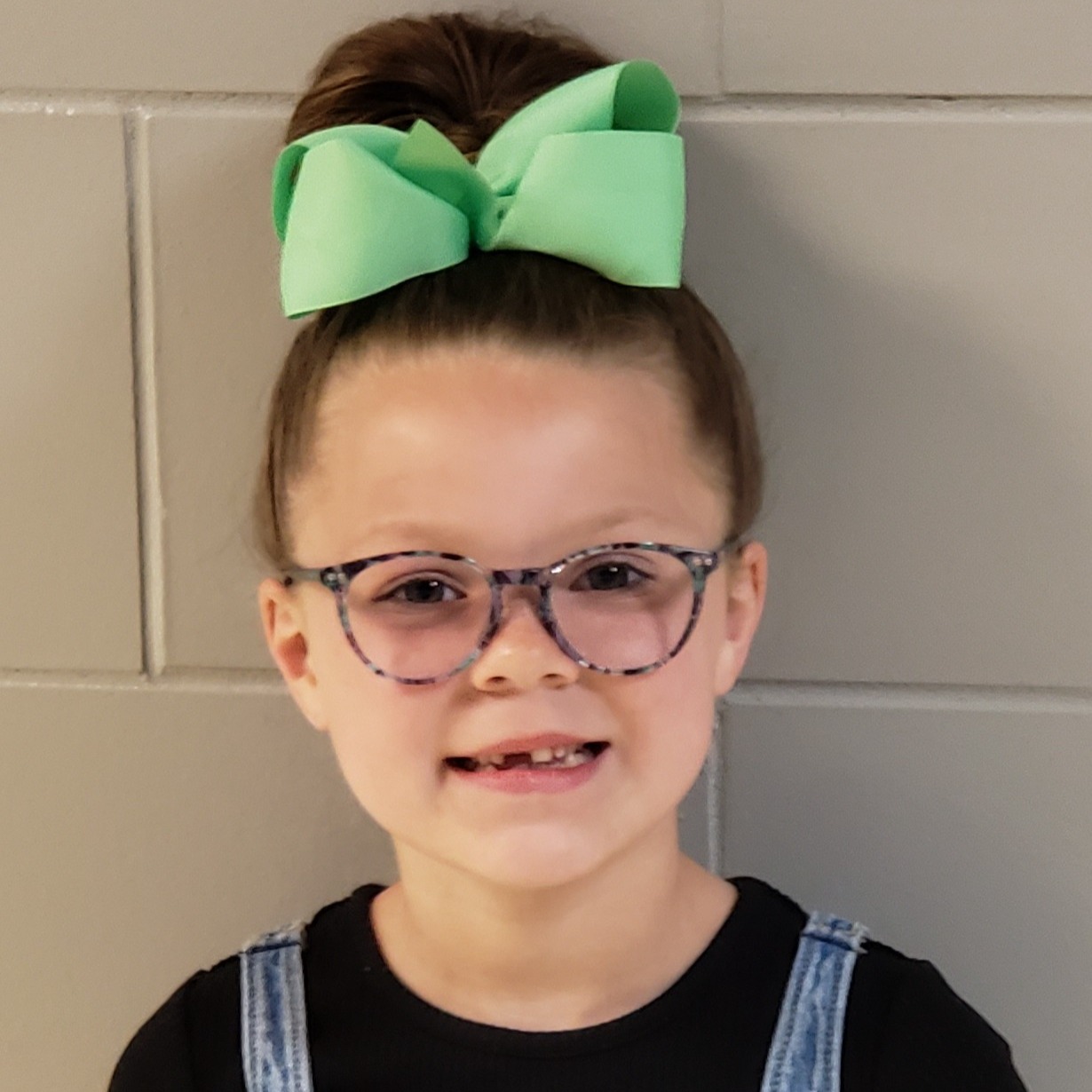 smiling girl with dark hair pulled up in a messy bun with a big mint green bow and glasses missing a front tooth wearing a black t-shirt under an overall dress