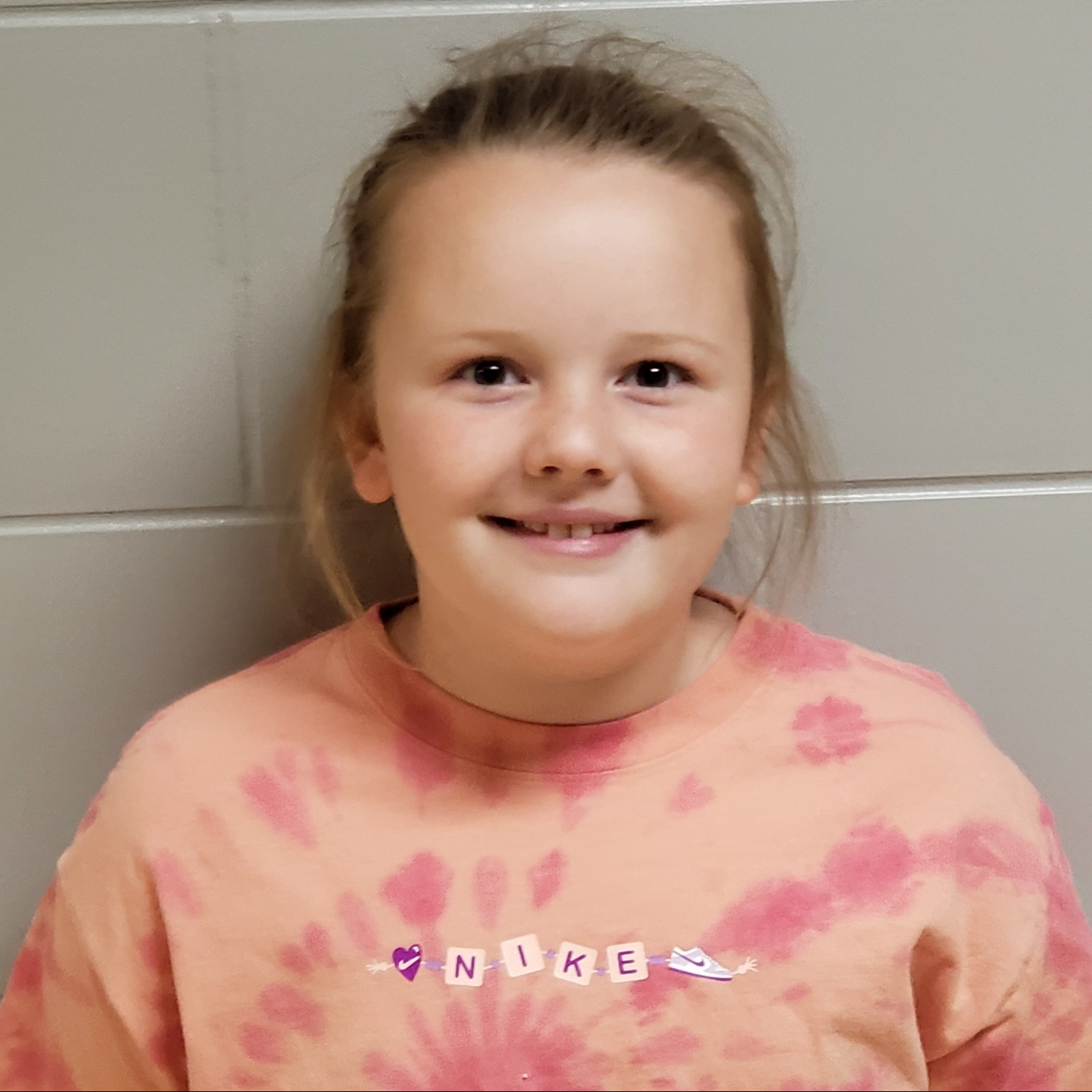 smiling young girl with dark blonde hair pulled back wearing a peach and pink tie-dyed t-shirt