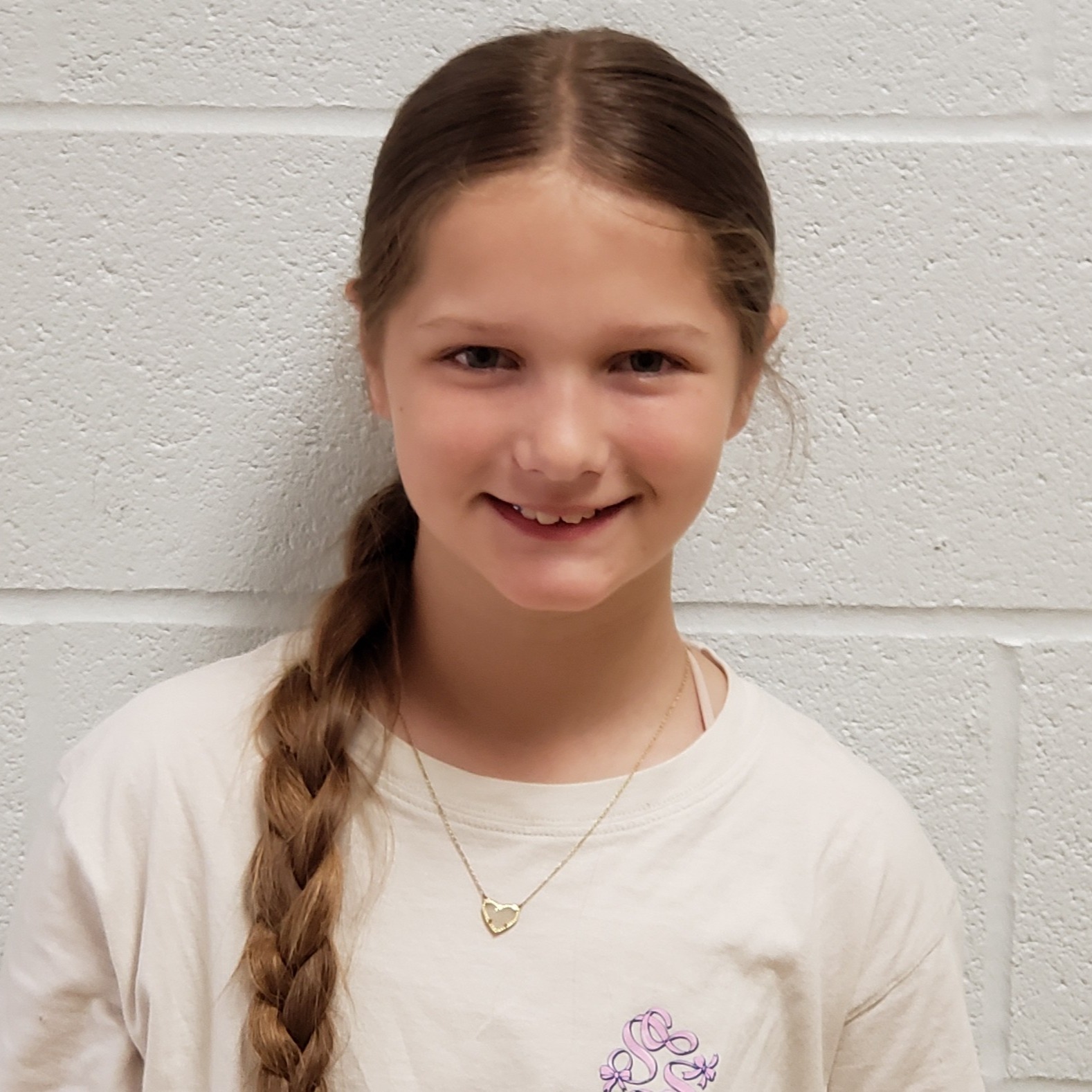 smiling girl with long brown hair pulled back into a side braid wearing a white t-shirt and gold heart necklace