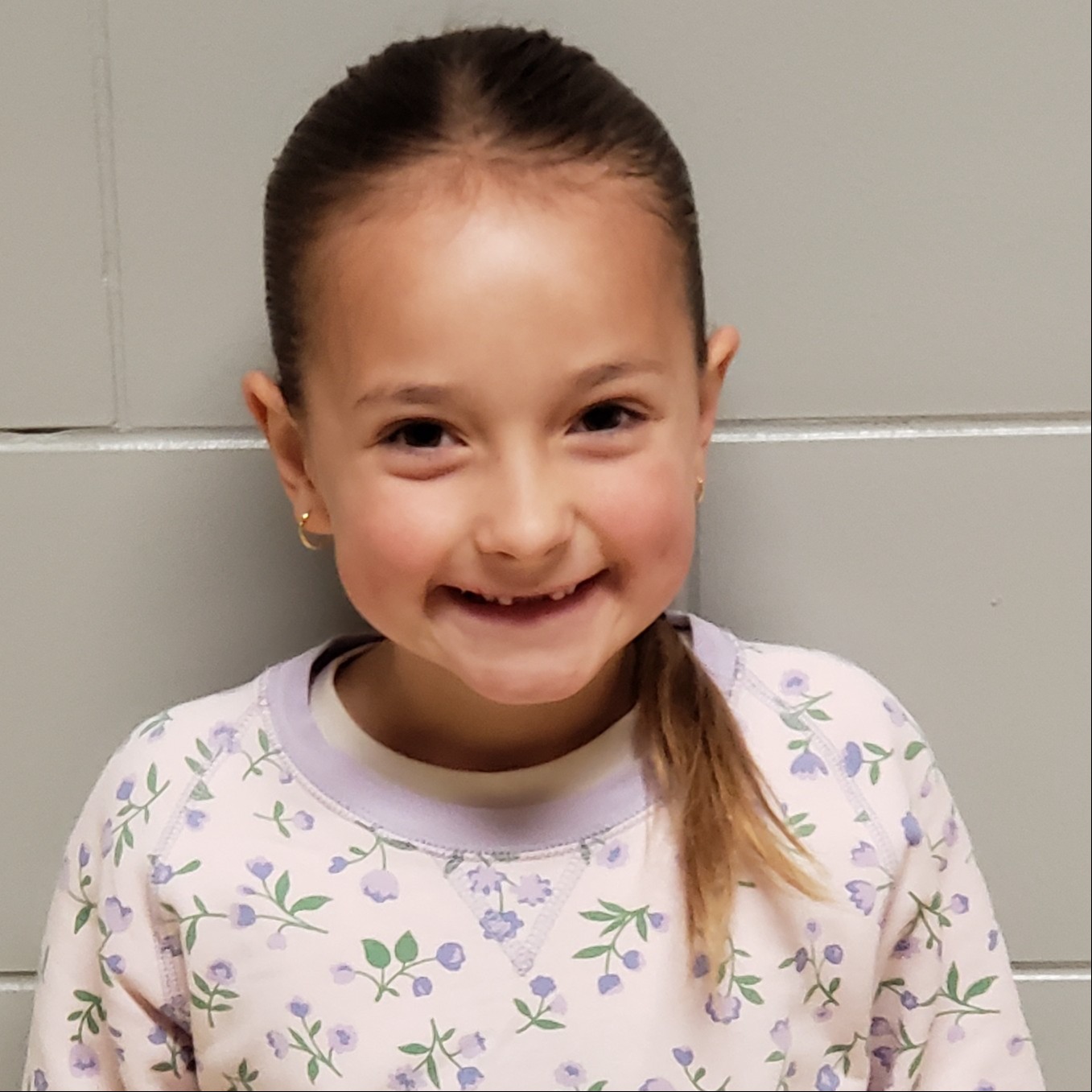 young girl with dark hair pulled back and missing front teeth wearing a light purple sweatshirt with purple flowers on it