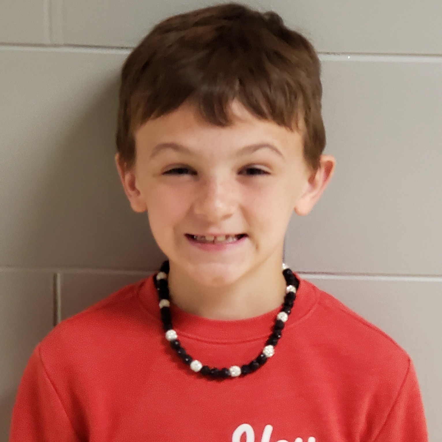 smiling  boy with reddish brown hair wearing a red t-shirt and a black & white beaded necklace