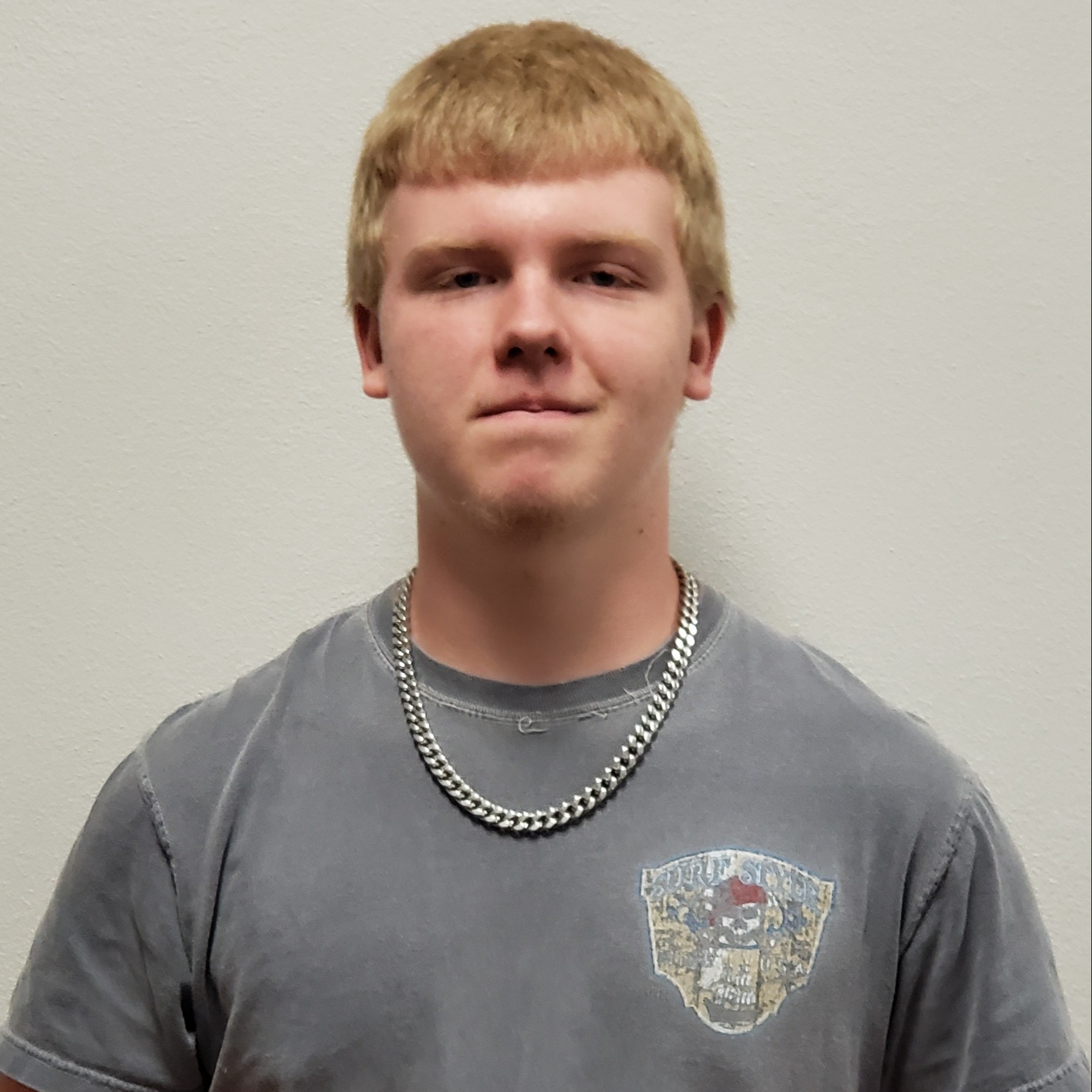 grinning young man with blonde hair wearing a gray t-shirt and silver chain 