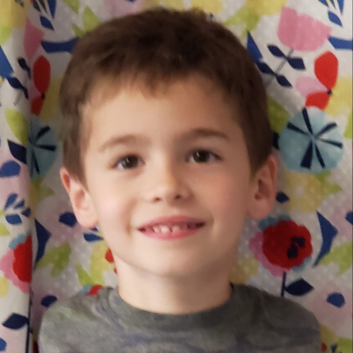 smiling little boy with dark hair wearing a camouflage t-shirt, standing in front of a curtain with multicolored flowers on it