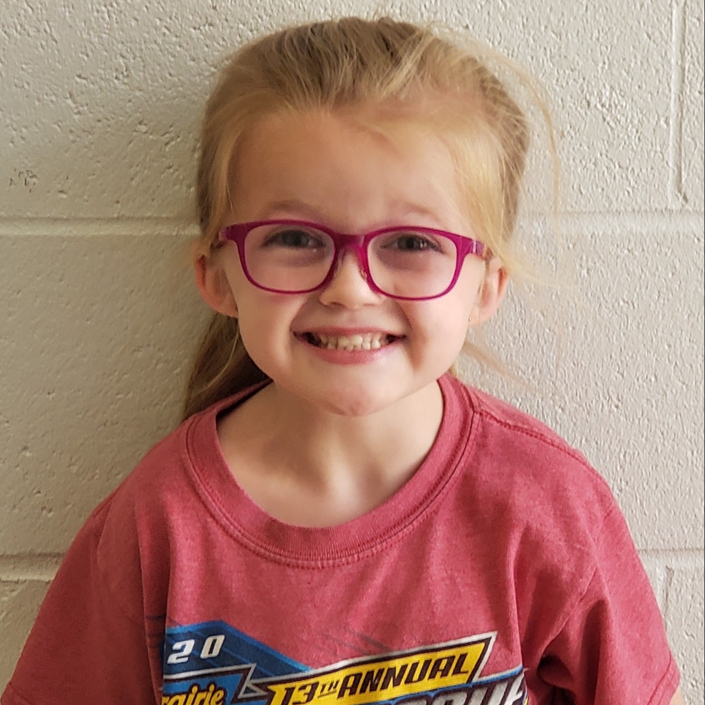 smiling little girl with blonde hair pulled back wearing a pink t-shirt and glasses