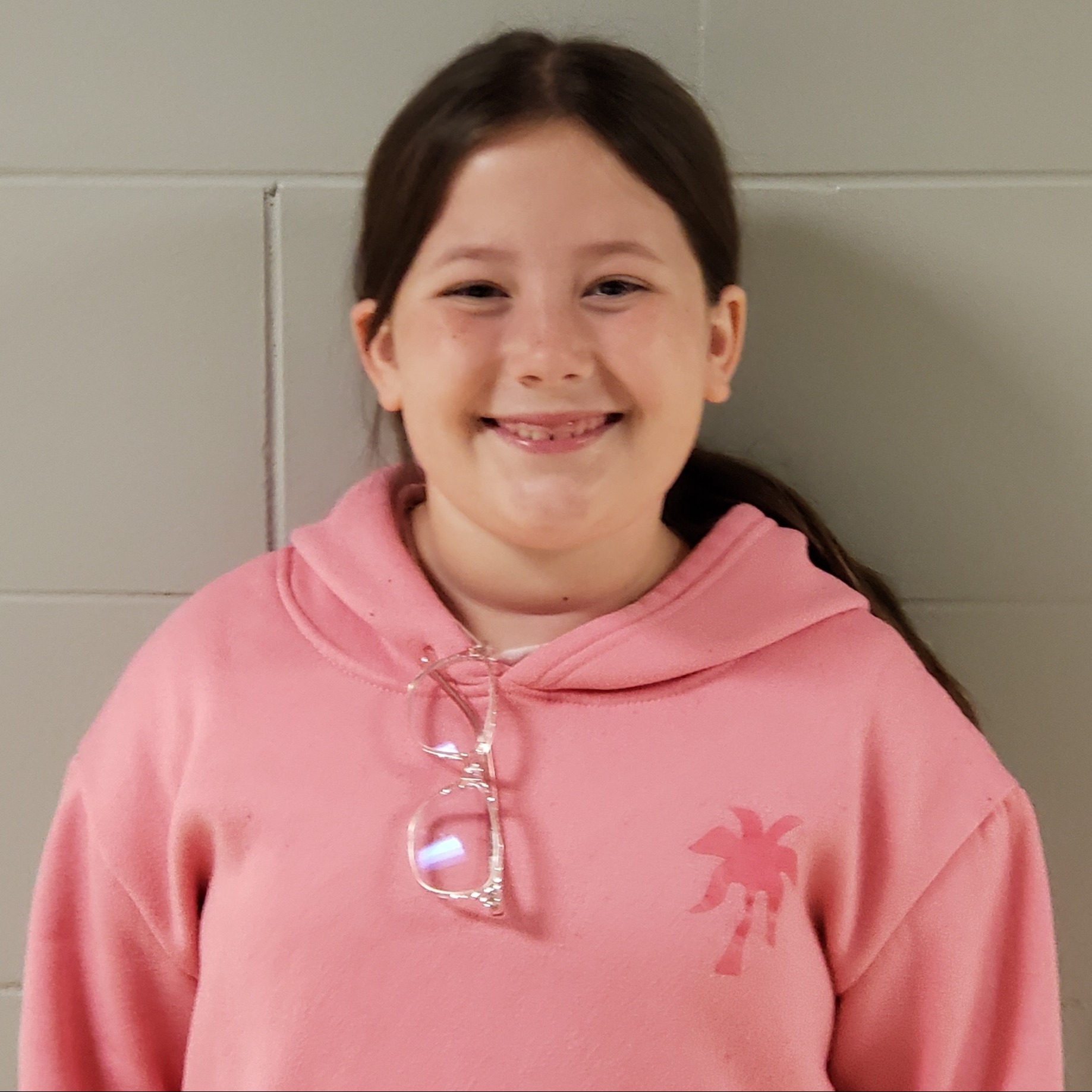 smiling girl with dark hair pulled back wearing a pink hoodie with her glasses hanging from the neck