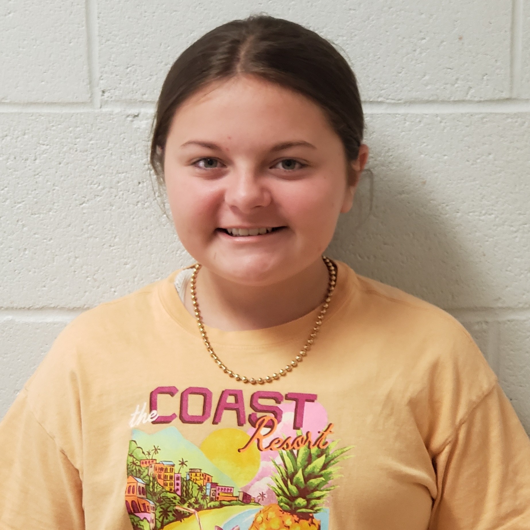 smiling girl with brown hair pulled back wearing a yellow Coast Resort t-shirt and gold beads