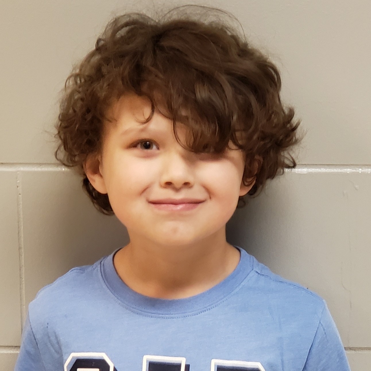 grinning young boy with dark curly hair hanging over one eye wearing a light blue t-shirt