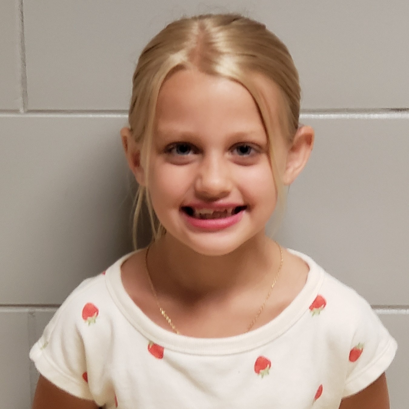 smiling young girl with blonde hair pulled back wearing a white t-shirt with strawberries and bright pink lipstick