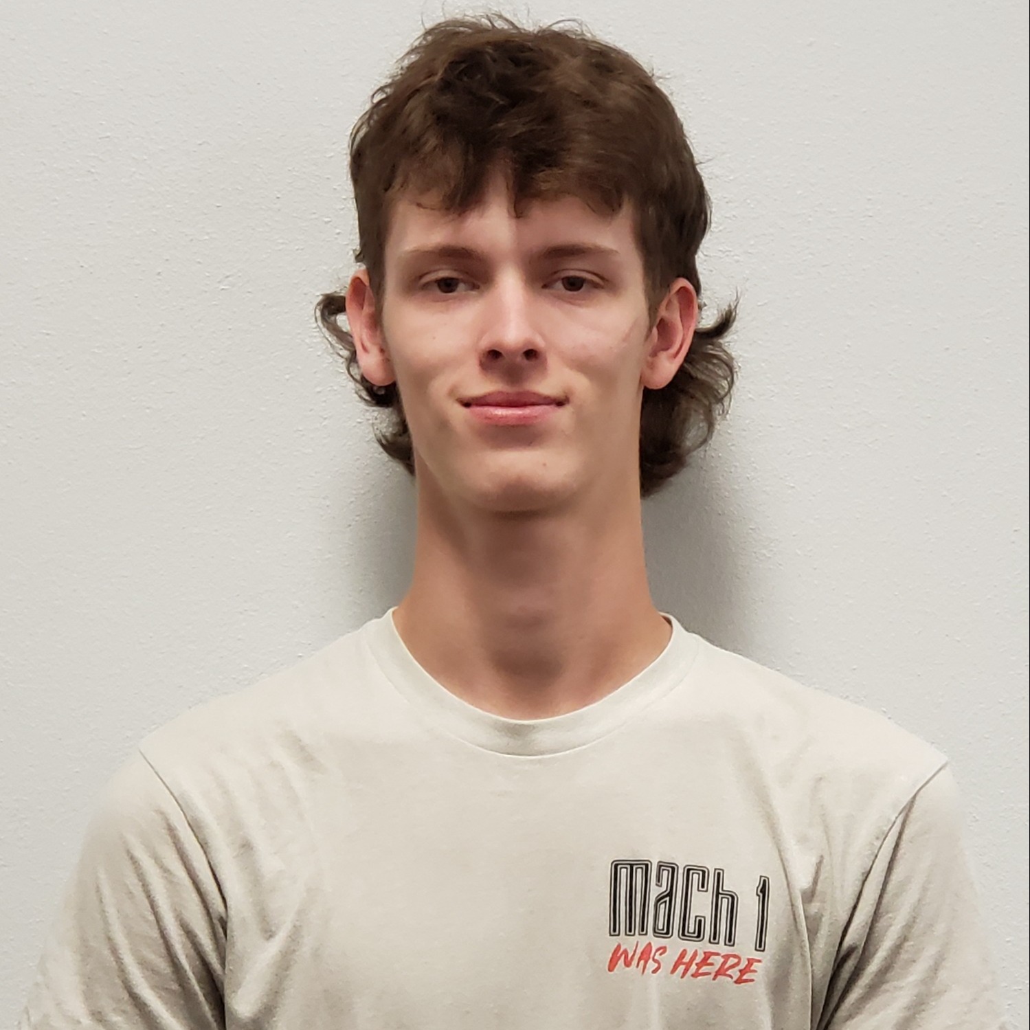 grinning young man with a brown mullet hairstyle wearing a tan Mach 1 t-shirt