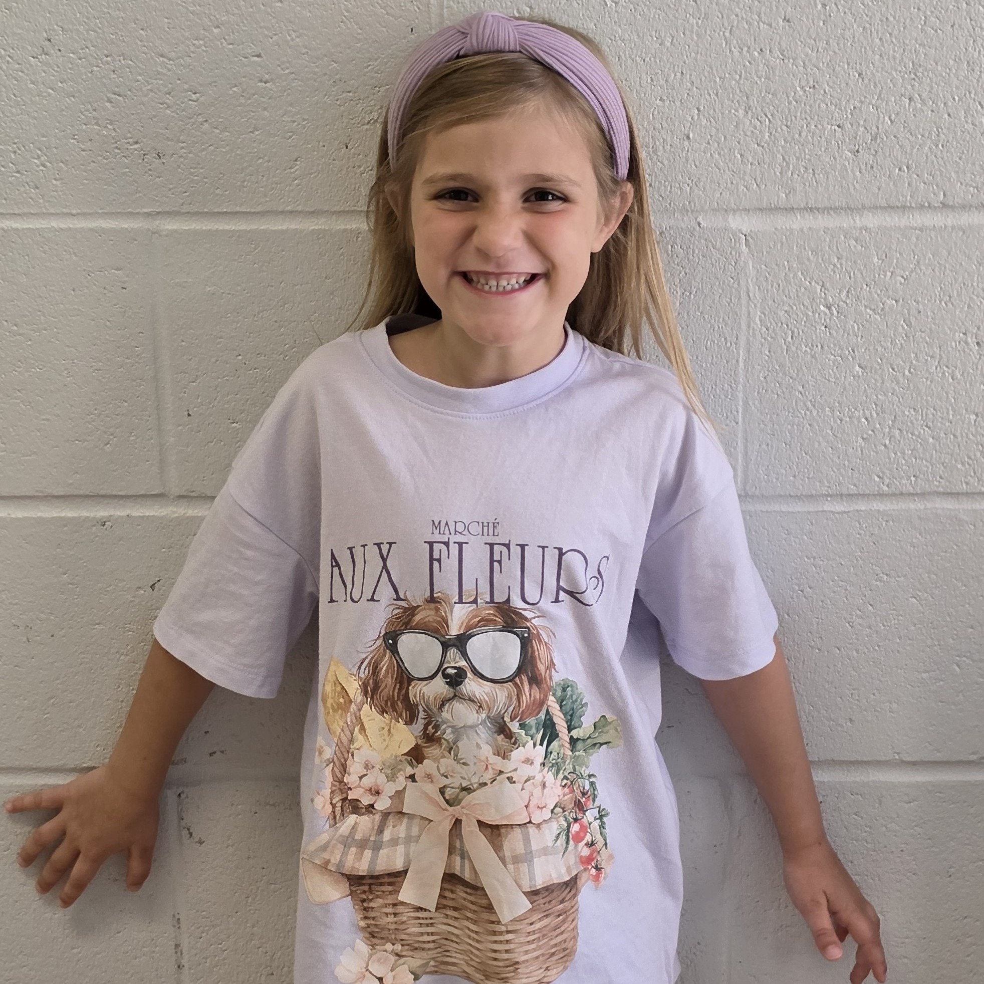 smiling girl with sandy blonde hair wearing a lilac t-shirt with a puppy in sunglasses on it and a lilac headband