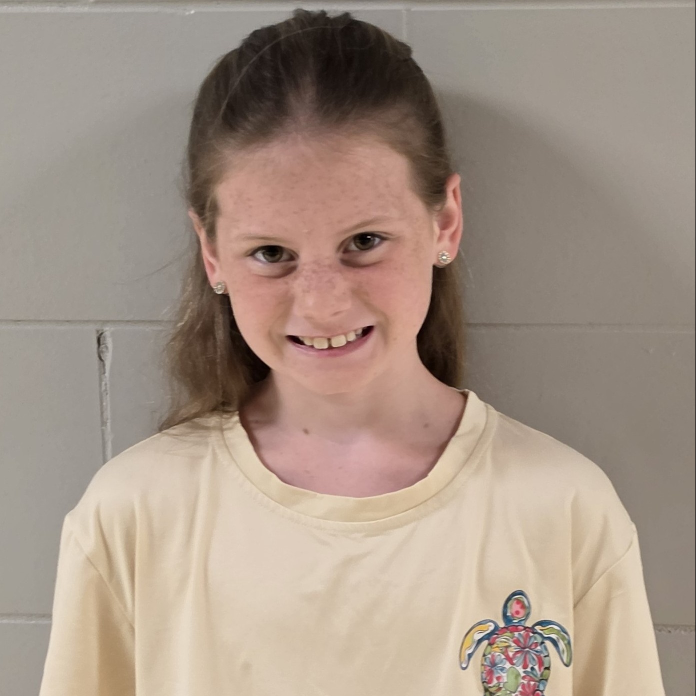 smiling girl with reddish brown hair pulled back and freckles wearing a light yellow t-shirt 