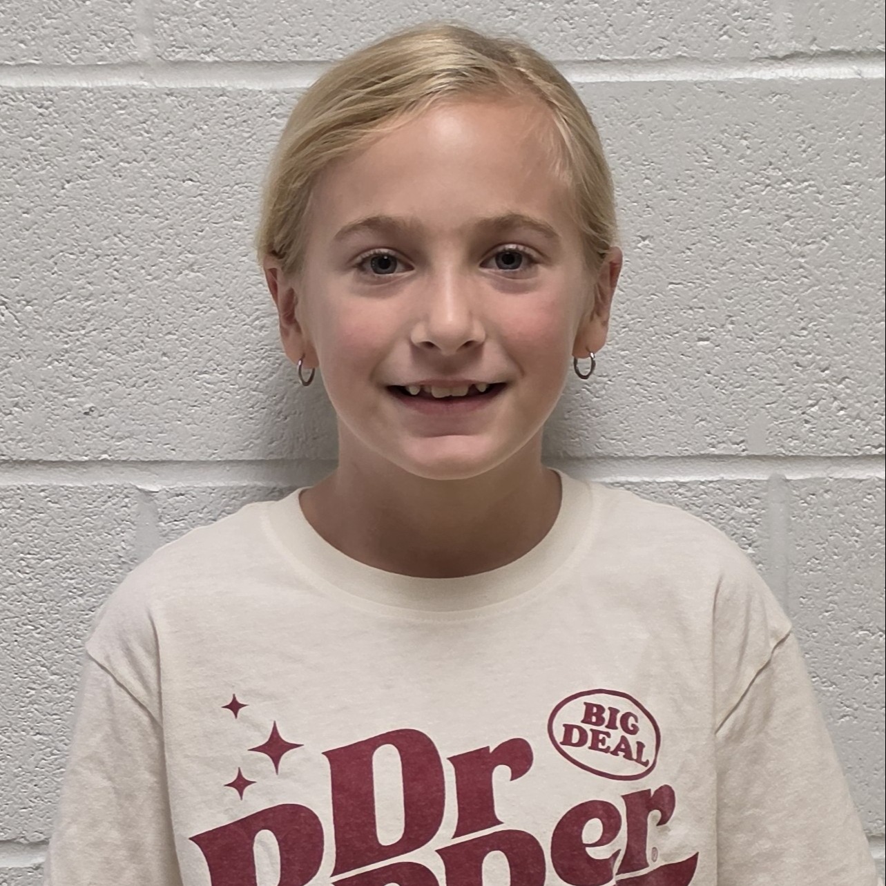 smiling girl with blonde hair pulled back and hoop earrings wearing a cream Dr Pepper t-shirt