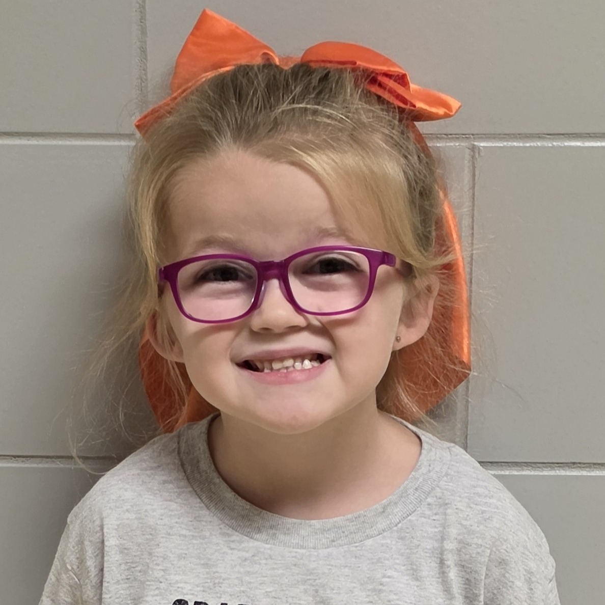smiling young girl with blonde hair pulled back in an orange bow wearing pink glasses and a gray t-shirt