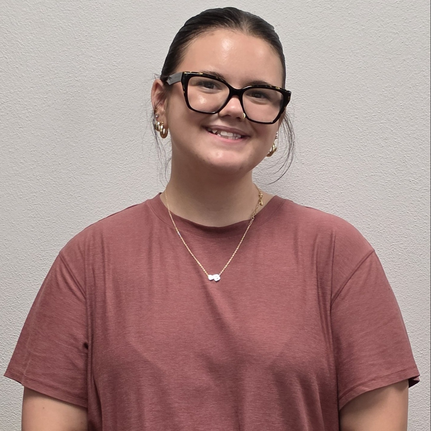 smiling young lady with dark hair pulled back, multiple ear piercings and a nose piercing wearing a light red t-shirt, dark-rimmed glasses, and a gold necklace