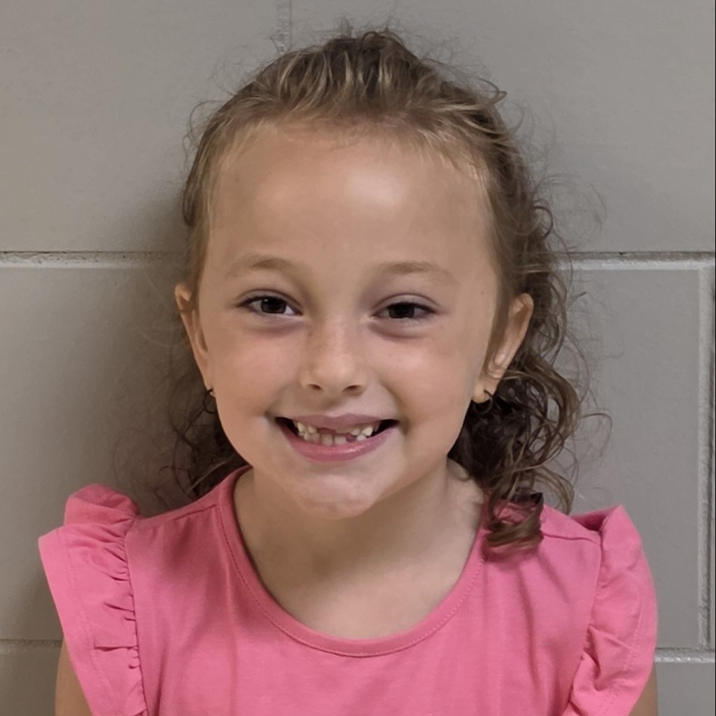 smiling girl with brwn curly hair pulled half up wearing a pink ruffle sleeve t-shirt