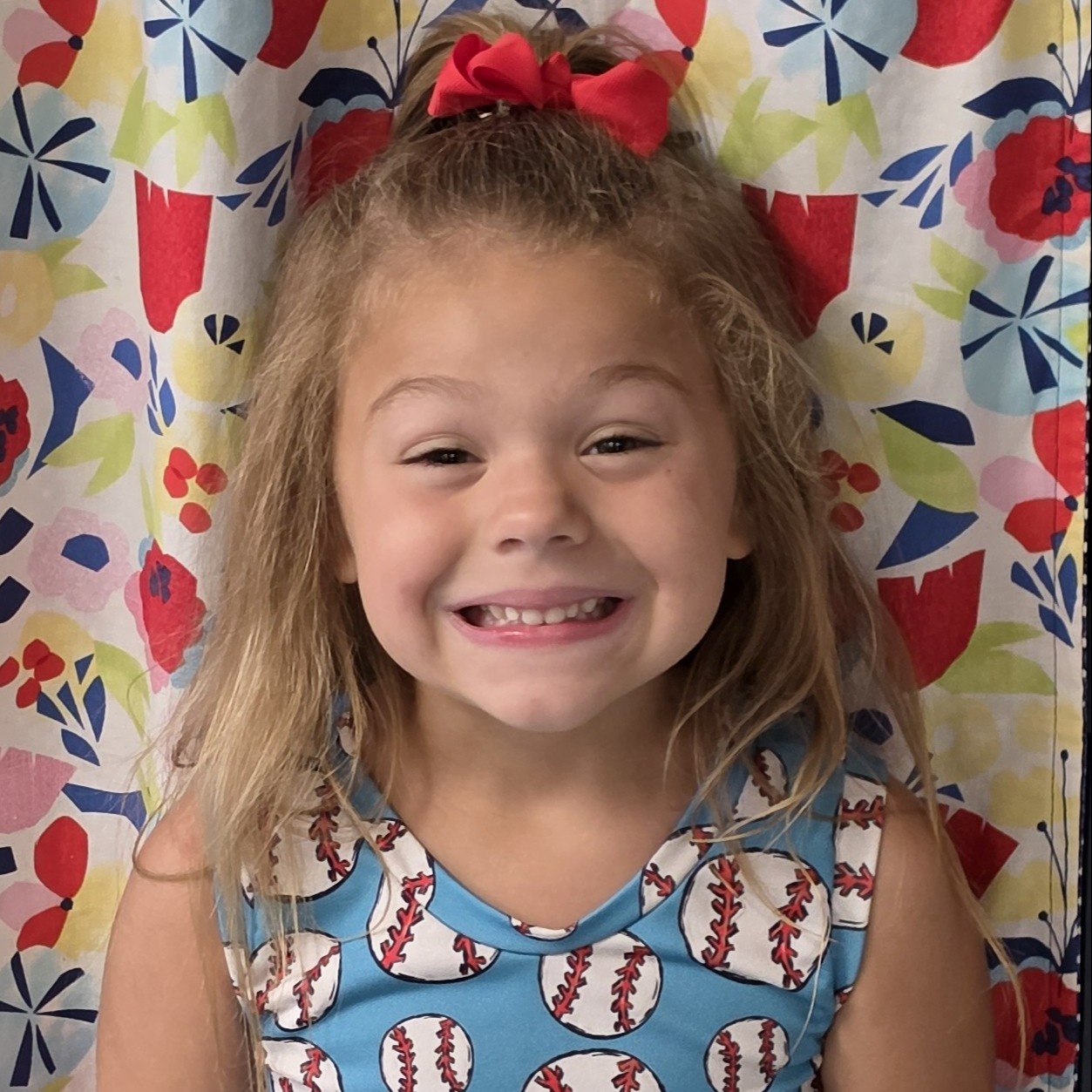 smiling little girl with light brown hair pulled half up with a red bow wearing a light blue tank top with baseballs all over it