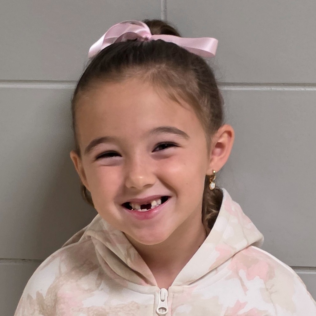 smiling girl with brown hair pulled up in a pink bow and missing front teeth wearing a pink hooded sweatshirt zipped up