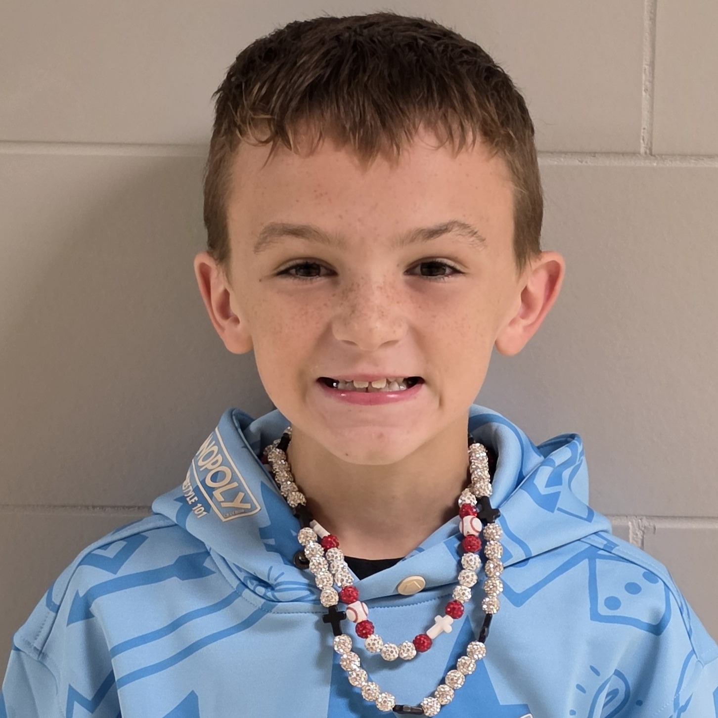 smiling boy with brown hair and freckles wearing a blue hooded sweatshirt and sparkly necklace with baseballs and crosses