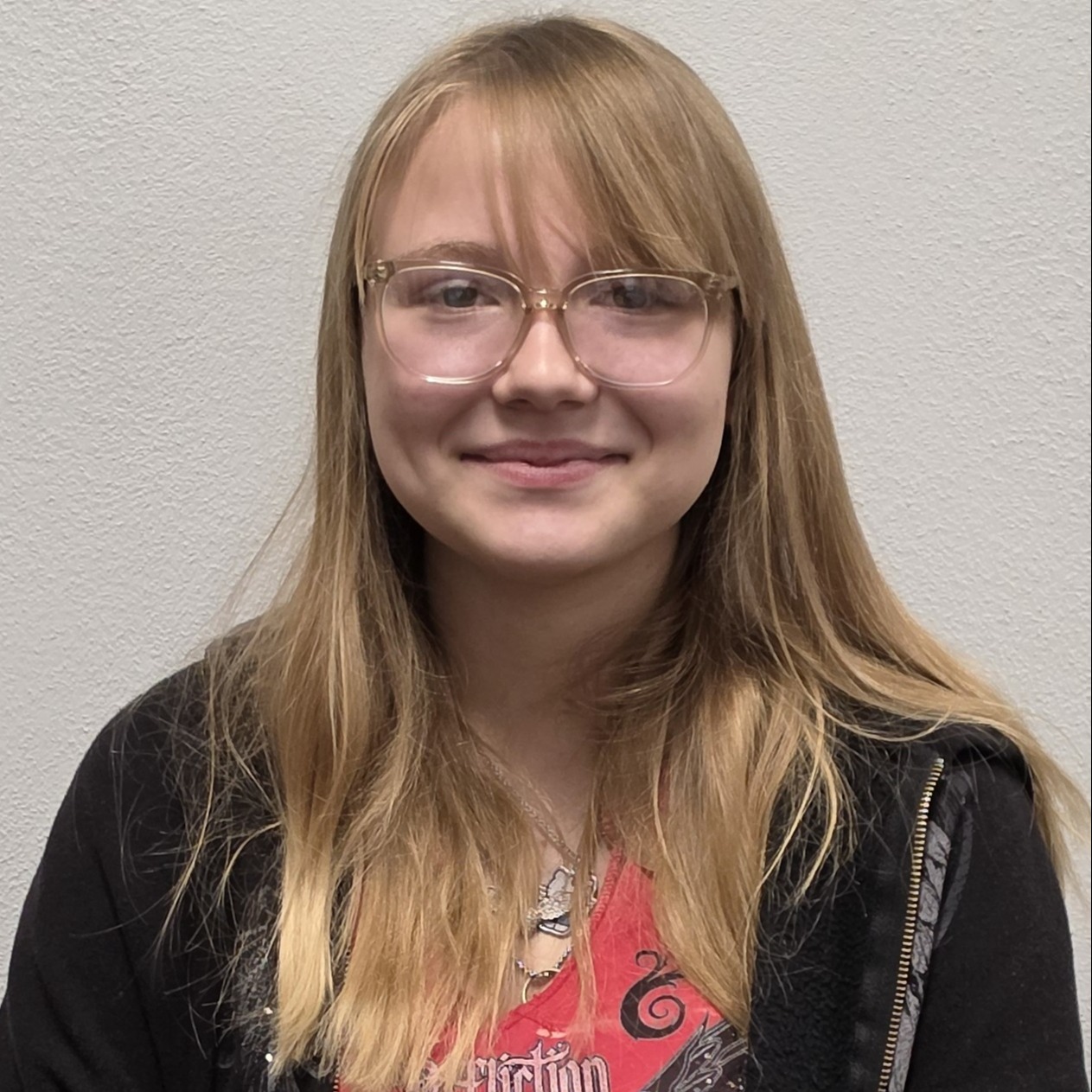 grinning girl with sandy blonde  hair and glasses wearing a red t-shirt under a black jacket