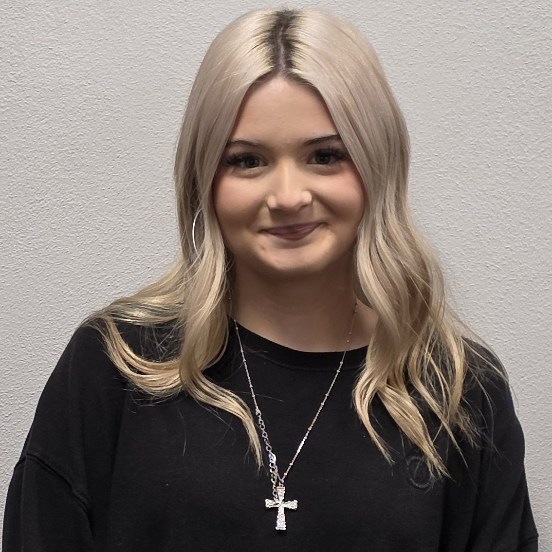 grinning young lady with long, blonde hair wearing a black sweatshirt and silver cross necklace