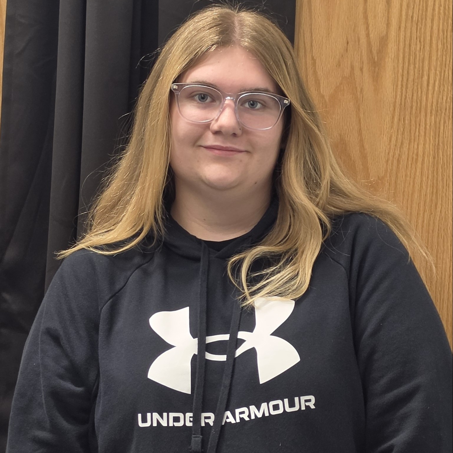 grinning young lady with long, blonde hair and glasses wearing a black Under Armour hoodie