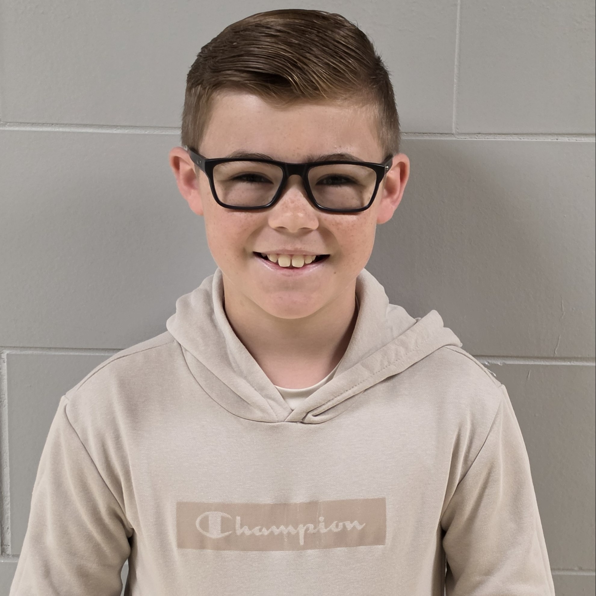 smiling boy with brown hair and black-rimmed glasses wearing a tan hooded sweatshirt