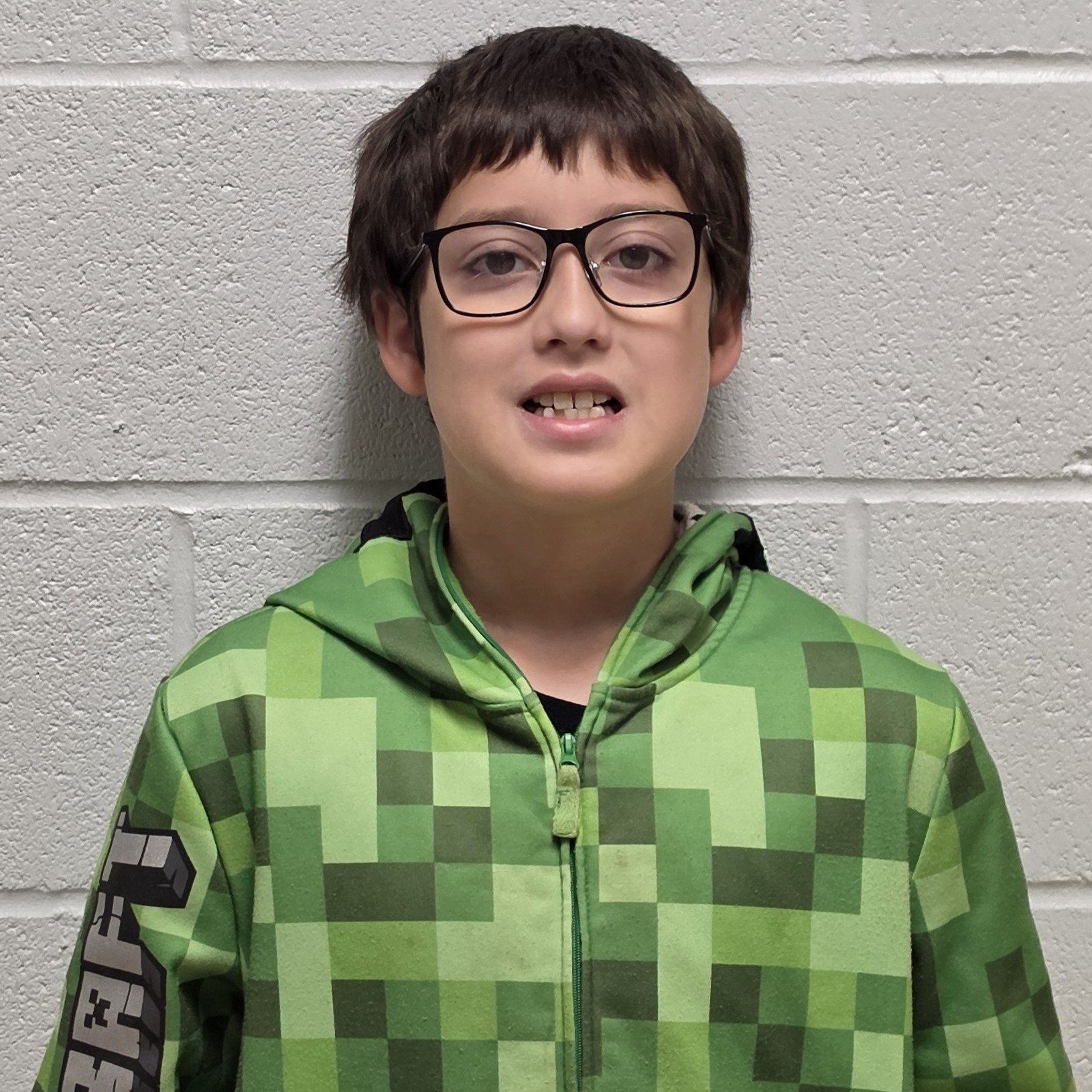 smiling boy with brown hair and black-rimmed glasses wearing a greenjacket zipped up over a black t-shirt