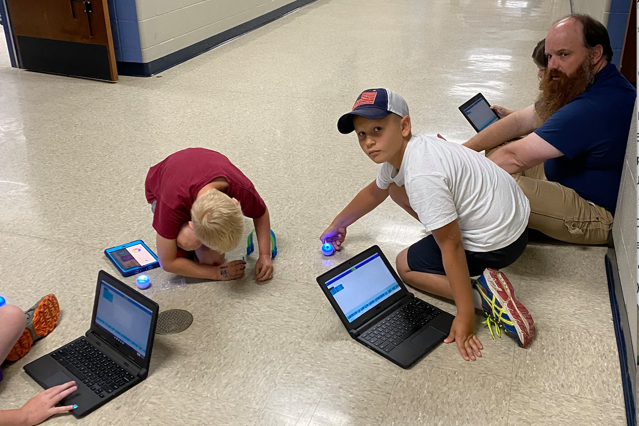 kids sitting in floor using laptops and mini-robots 