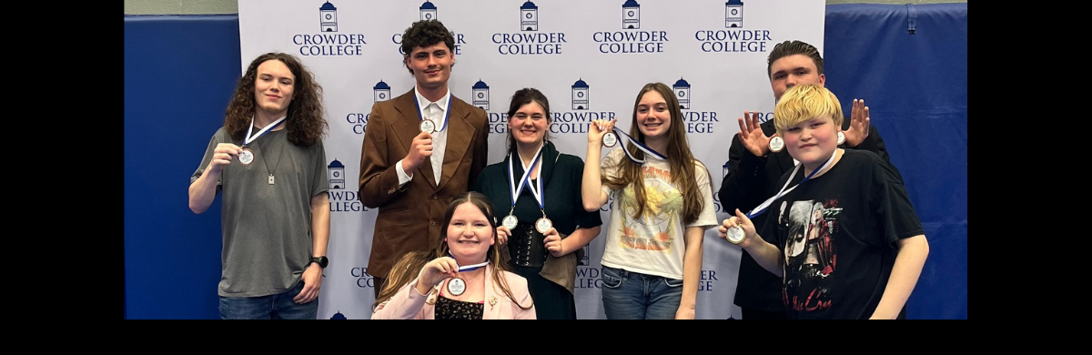 A wide, landscape-oriented group photograph of seven students posing with medals in front of a professional backdrop. The white backdrop features a repeating "step and repeat" logo pattern for "Crowder College," accompanied by a small blue bell tower icon.  The Subjects: Seven young adults (five standing and two seated or kneeling in the front) are arranged for a group portrait. They appear to be celebrating a "Business Contest" achievement.  The Medals: Each student is holding up or wearing a circular medal attached to a blue and white ribbon.  The Background: The white Crowder College backdrop is flanked on both the far left and far right by solid blue curtains or banners.  Clothing &amp; Style: The students are dressed in a mix of professional and casual attire:  One student wears a brown suit jacket.  Others wear t-shirts (including a black graphic tee), sweaters, and jeans.  The overall mood is proud and celebratory.