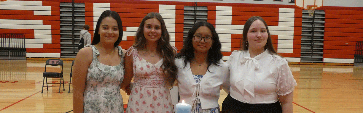 A horizontal group photograph of four young women standing side-by-side behind a candle-lighting display. They are posed in a school gymnasium for a National Honor Society (NHS) induction ceremony for 2026.