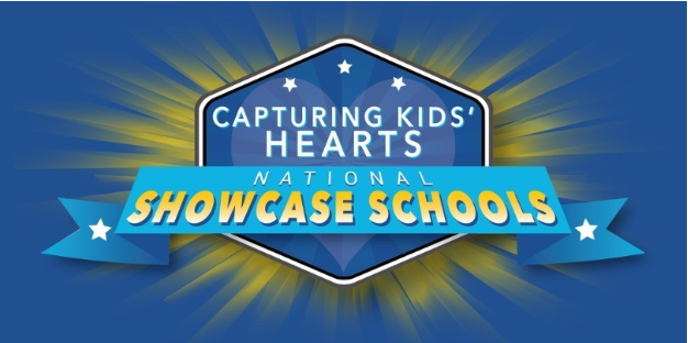A graphic logo for Capturing Kids’ Hearts National Showcase Schools. It features a dark blue hexagonal shield with three white stars at the top and the text "CAPTURING KIDS' HEARTS" in white. A bright blue ribbon banner across the middle reads "NATIONAL SHOWCASE SCHOOLS" in bold yellow and white lettering. The emblem is set against a dark blue background with yellow light rays radiating from behind the shield.