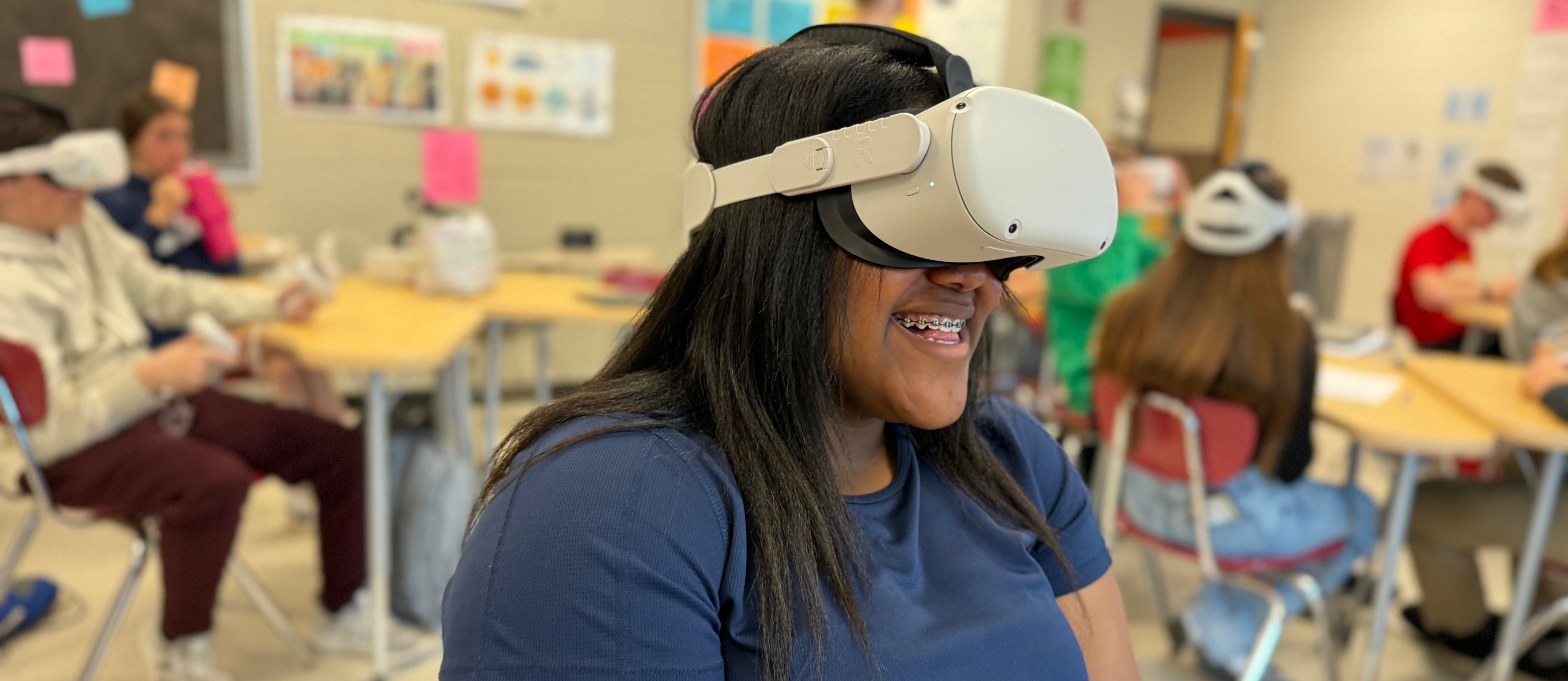 Female African American student with long straight black hair wearing VR headset and dark blue shortsleeve shirt is smiling. She has braces. 