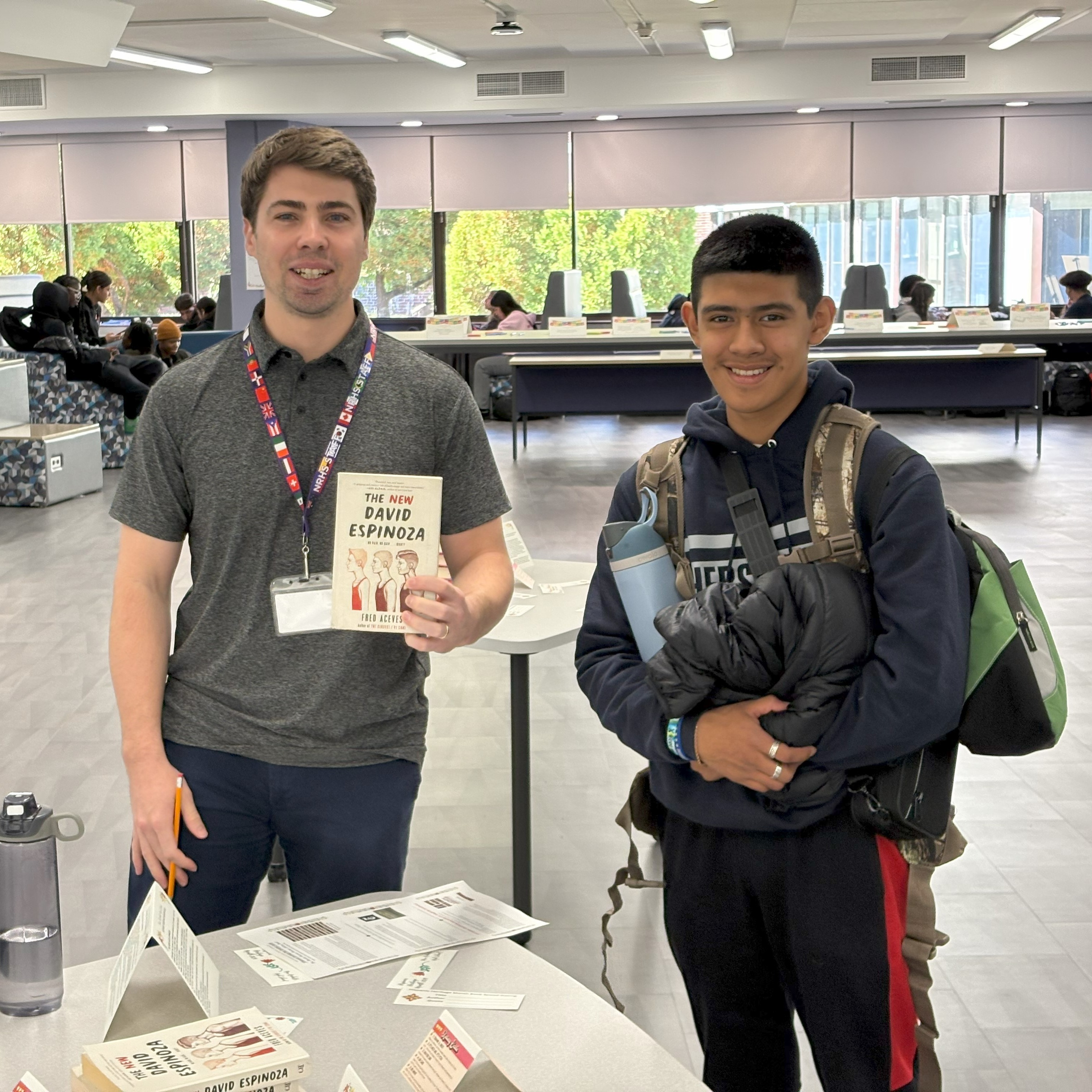 Mr. Beneville (English) stands with a student at a book tasting table, while holding a book.