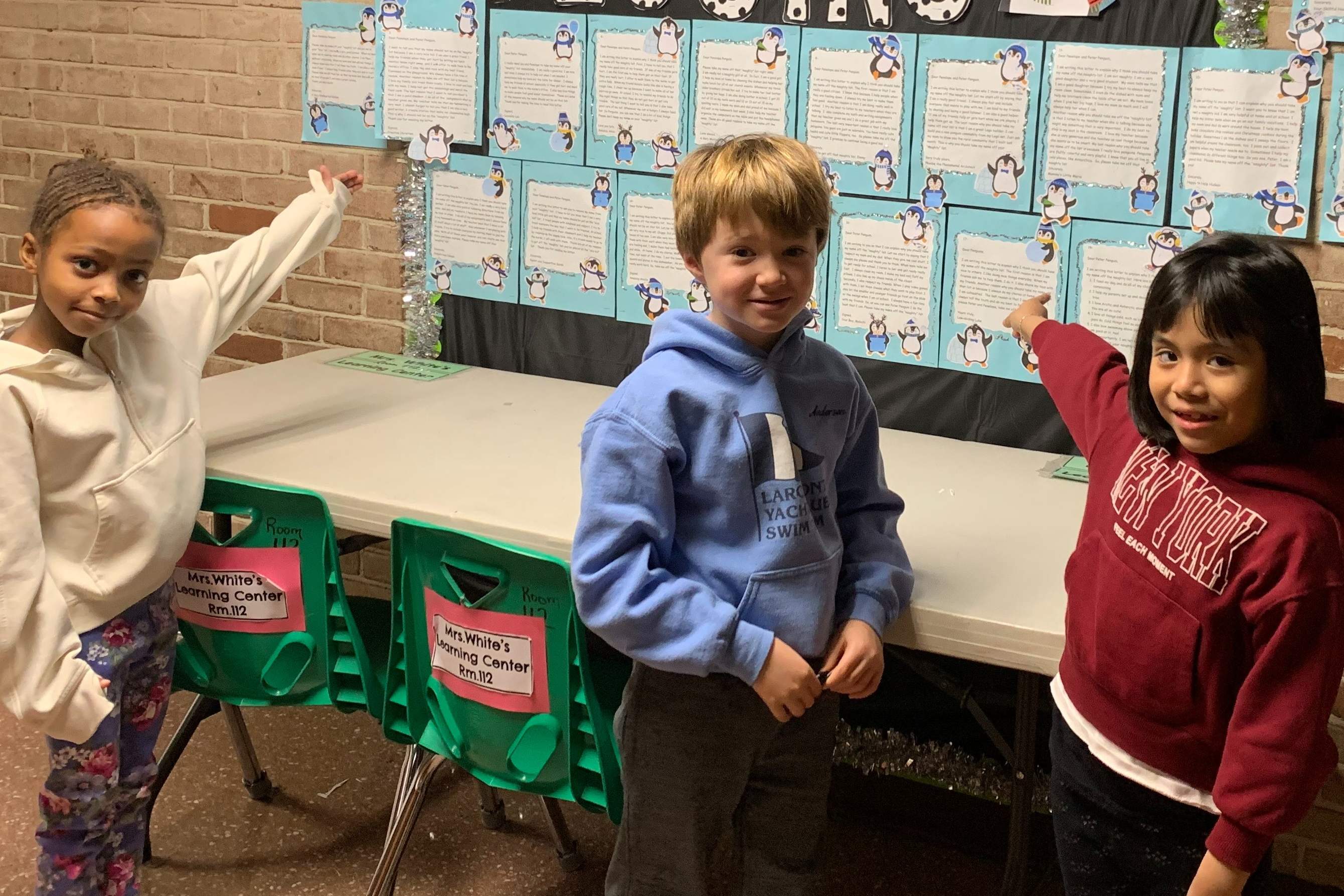 Three students point to their writing hanging on a bulletin board