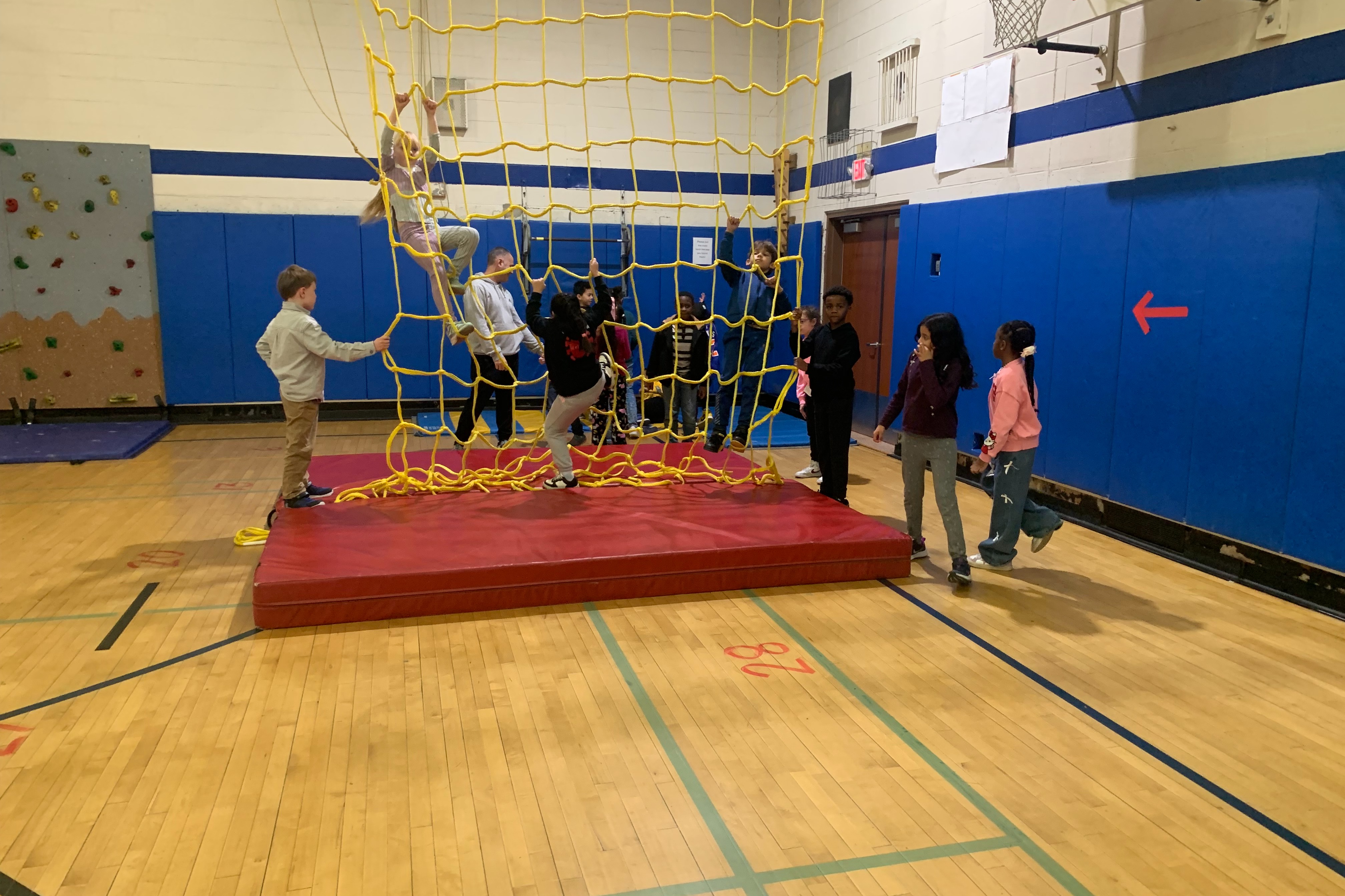 Physical Education Class students climb a yellow rope wall.