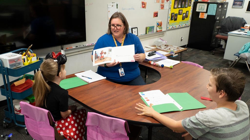 teacher sitting at a table holding up a book in front of two students