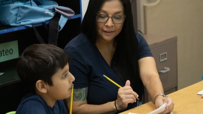 Teacher working with a student at a table, both holding pencils