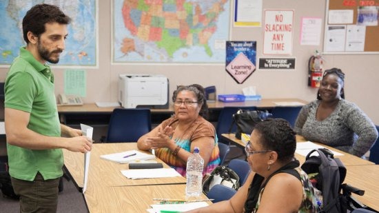 three adult students sitting at tables with teacher standing in the front