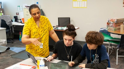 two students working with laptops and teacher standing next to them