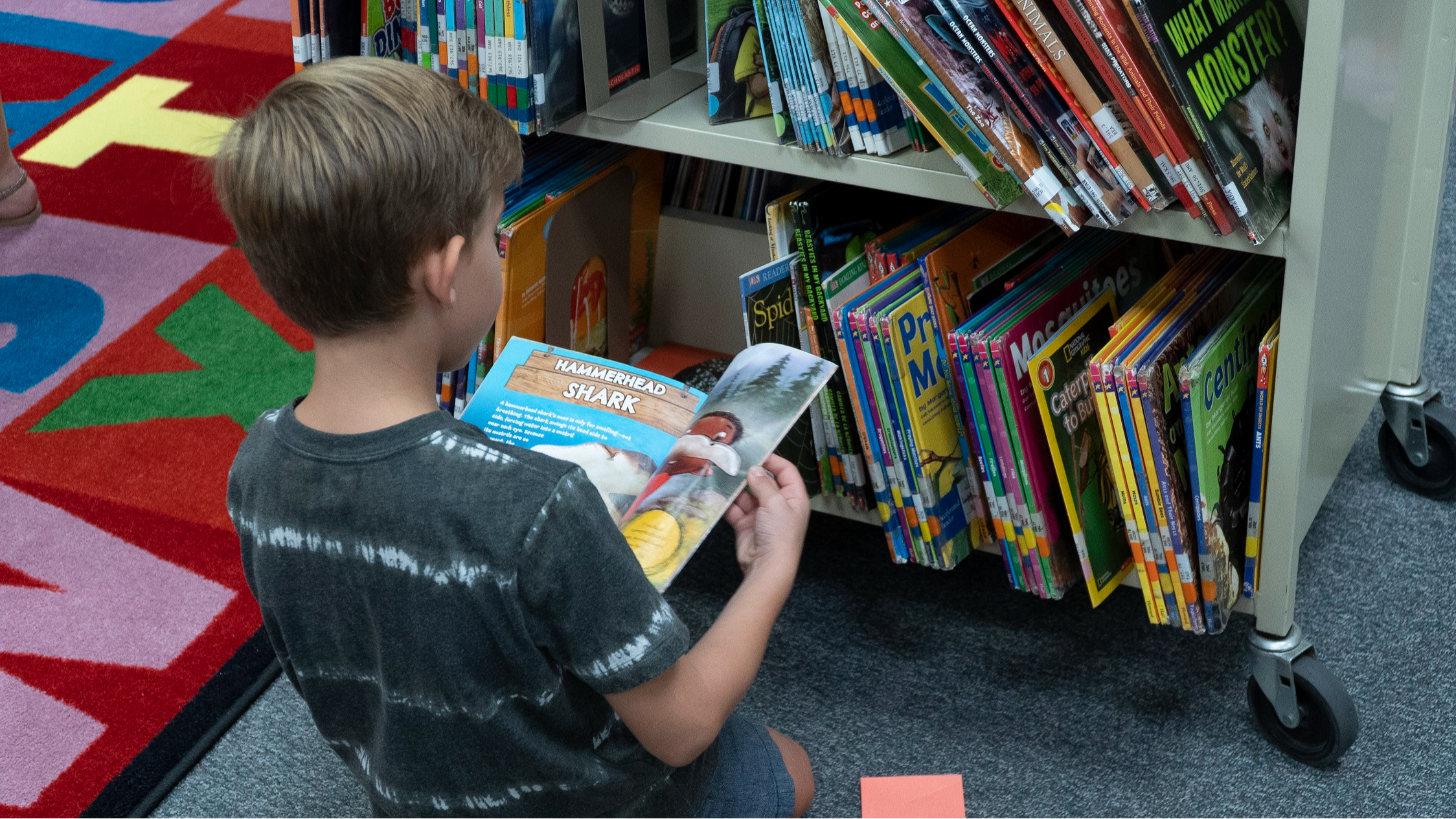 student sitting on floor in front of a book cart reading a book