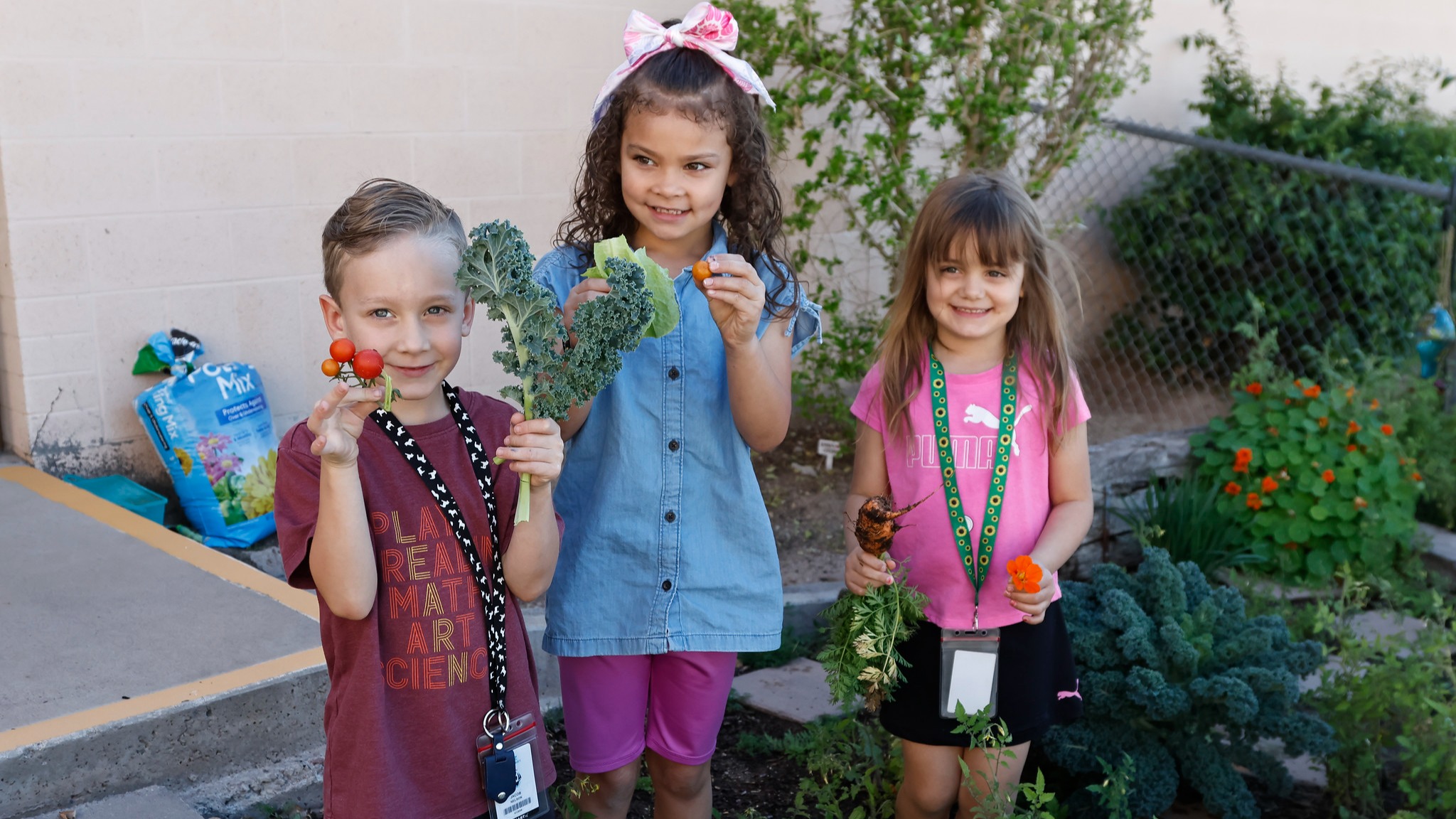 three students standing in a garden holdng vegetables