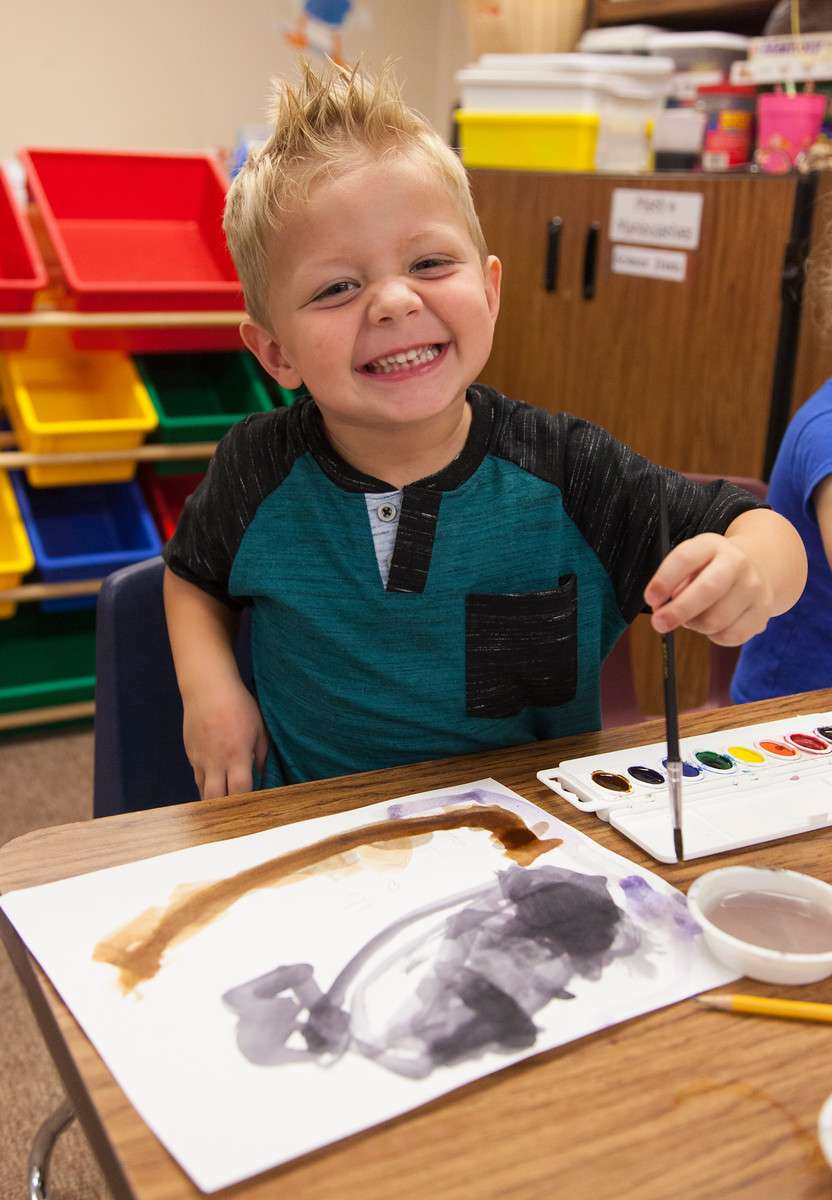 young boy painting in a classroom smiling