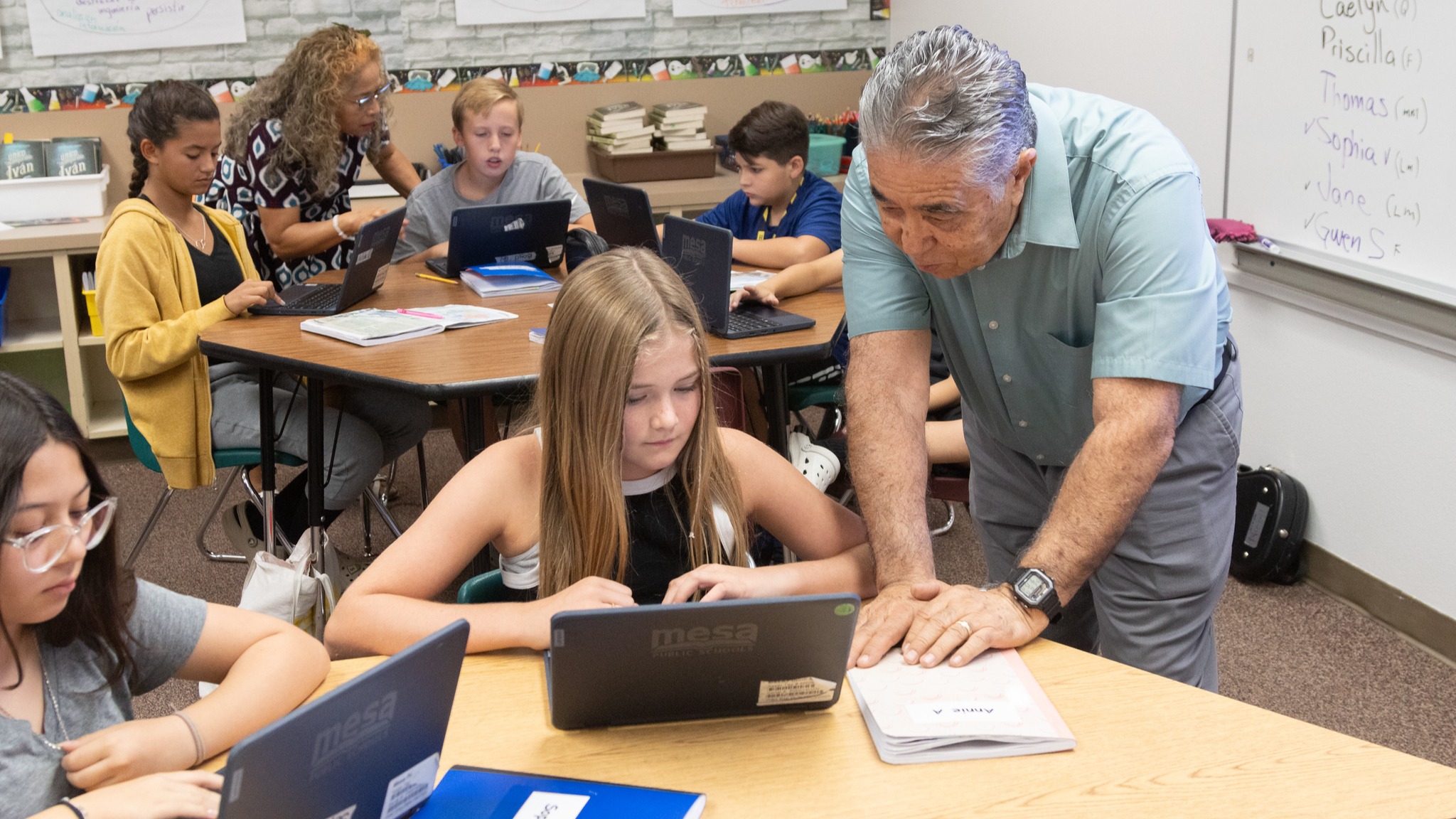 students sitting at tables working on laptops with teachers looking over them