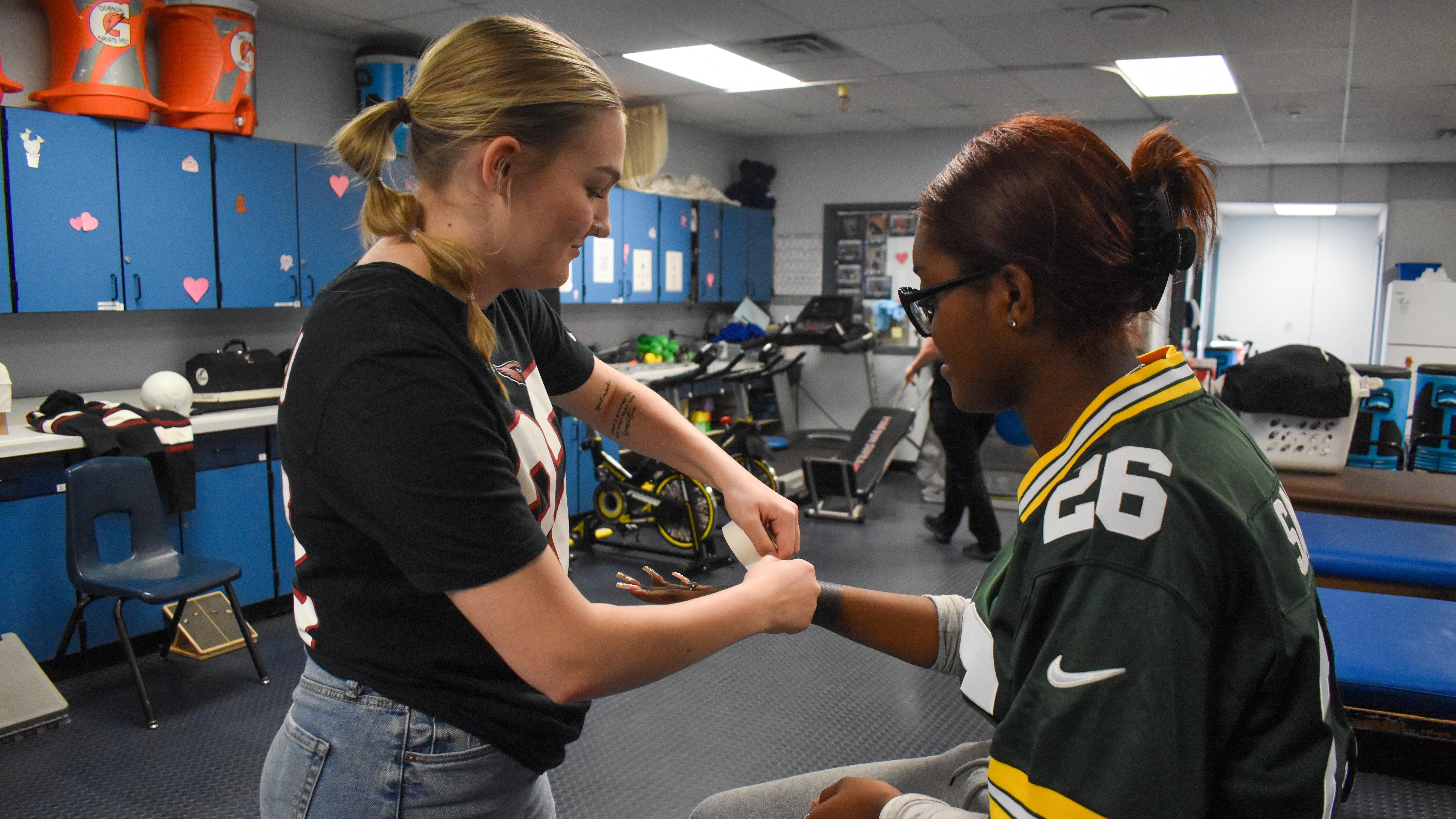 A student athletic trainer in a black shirt wraps the wrist of another student wearing a Green Bay Packers jersey in a school training room filled with exercise equipment.