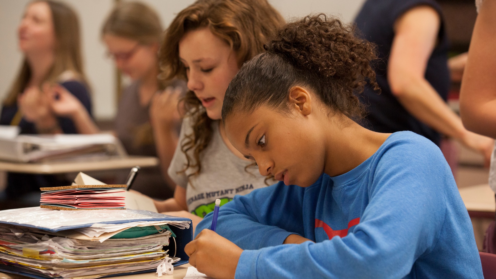 two students sitting at desks writing on paper with notebooks in front of them
