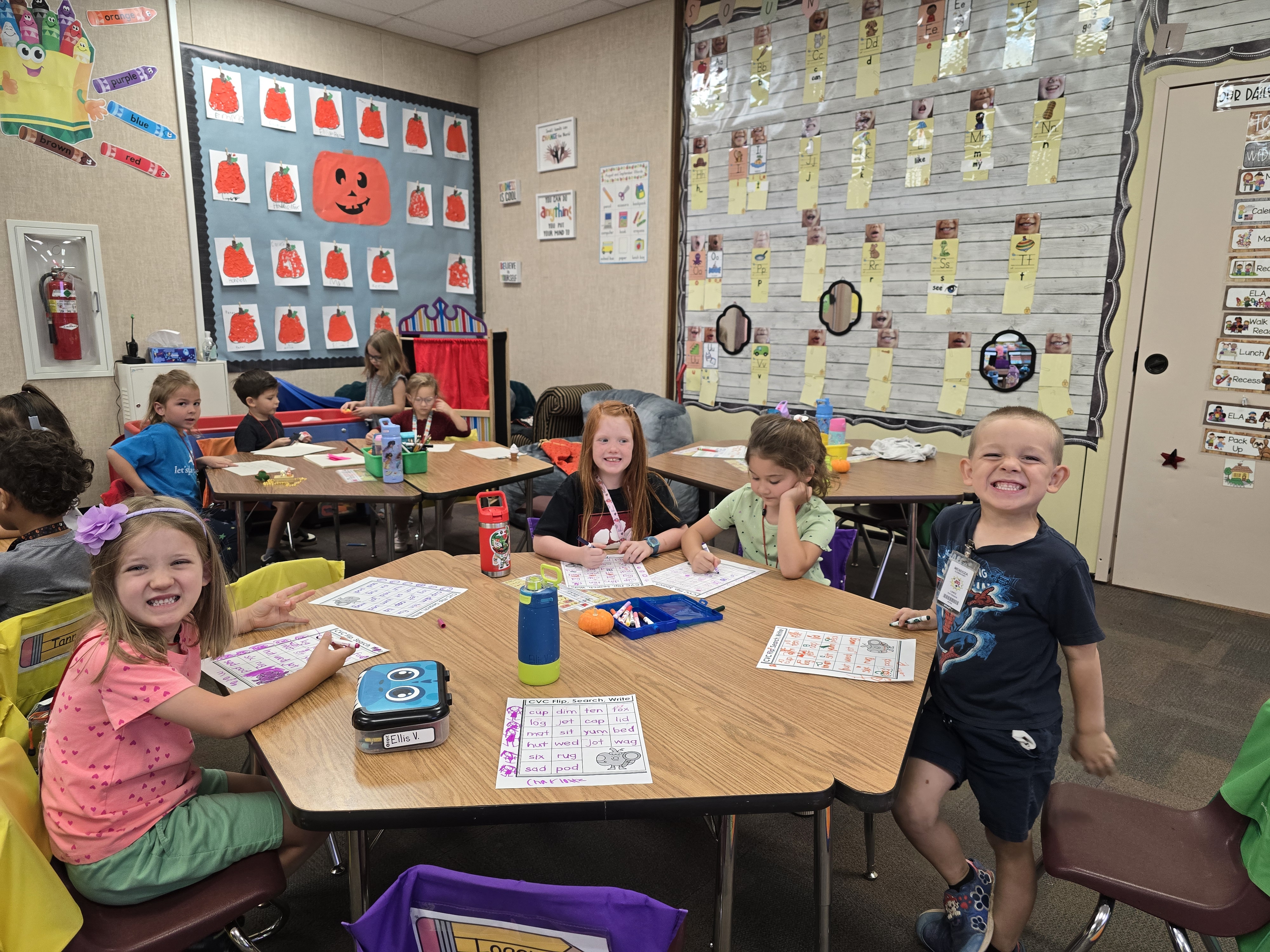 Several Kindergarten students sitting at their tables working on their school work and smiling for the camera