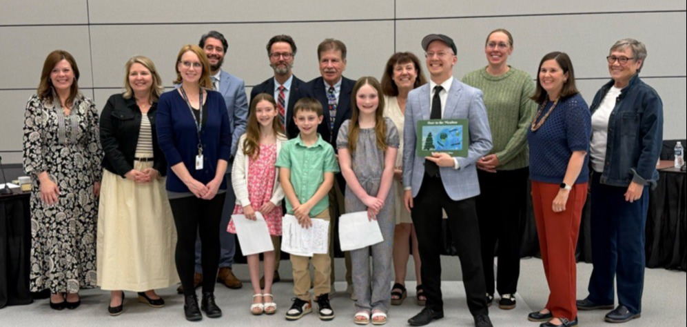 Several students surrounded by adults posing for a photo while a man holds up a book