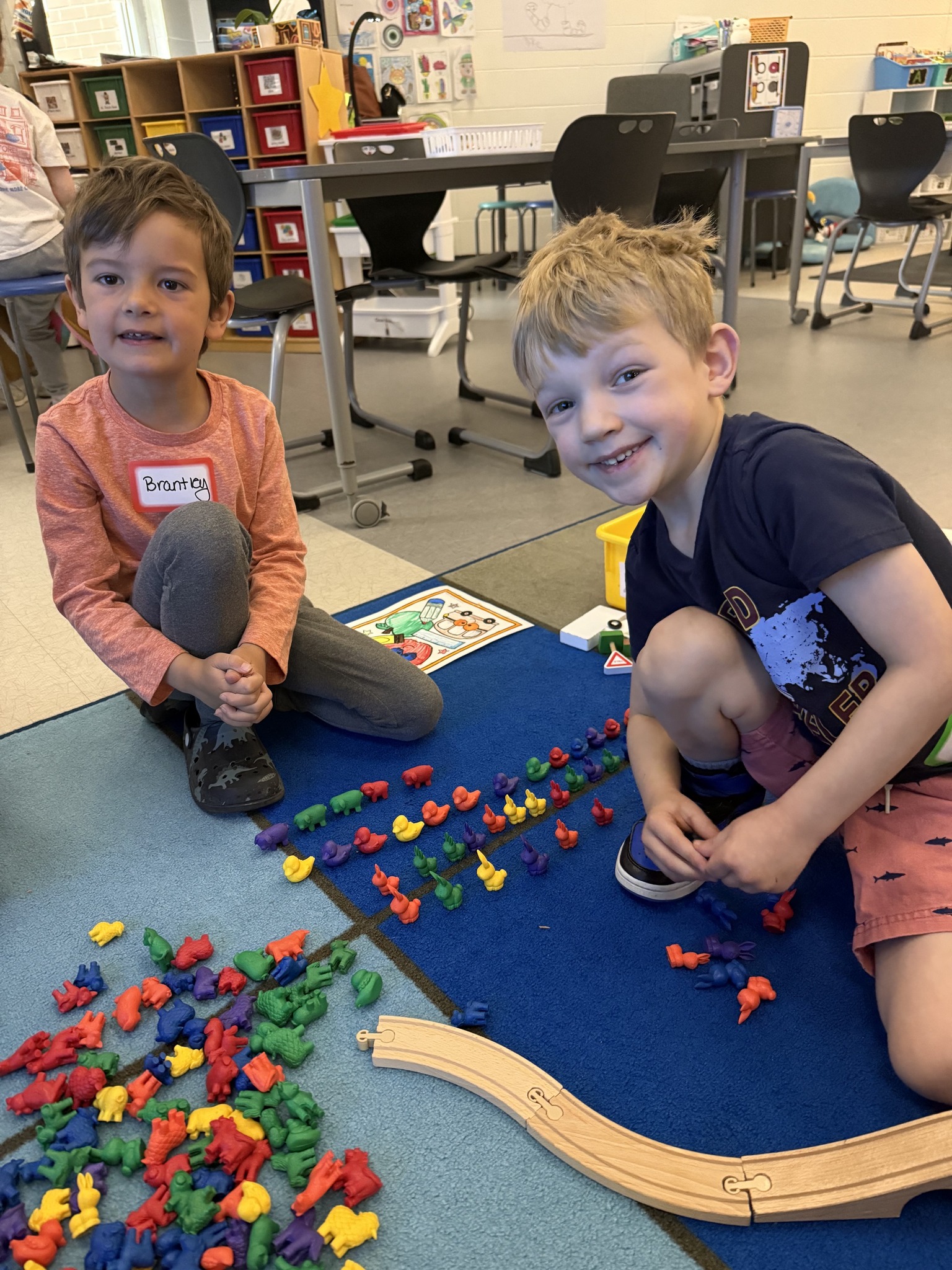two kids sitting on the floor smiling for a photo while in classroom